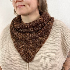 May include: A brown and tan crocheted cowl scarf. The triangular scarf is worn around the neck and has a textured, open weave design. The scarf is made of a soft, warm yarn and is a neutral accessory.