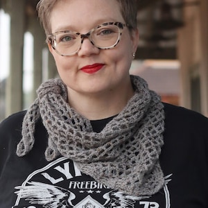 May include: A woman with short brown hair wearing a gray crocheted scarf and a black t-shirt with a graphic of wings and the text "FREEBIRD" on it.