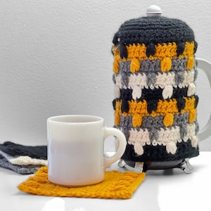 Pode incluir: Uma capa de cafeteira de crochê em preto, branco, cinza e amarelo com uma caneca de cerâmica branca e um descanso de copo de crochê amarelo.