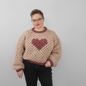 May include: A crocheted sweater with a large heart design in a darker shade of pink. The sweater is a light beige color and has a ribbed neckline and cuffs.