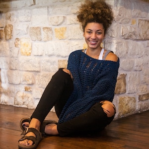 May include: A woman with curly brown hair is sitting on a wooden floor in front of a stone wall. She is wearing a blue knitted sweater, black jeans with holes, and brown sandals. She is smiling at the camera.