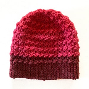 Peut inclure: Un bonnet en crochet avec un dégradé de couleurs rose et bordeaux. Le bonnet a un motif de point texturé et un bord replié.