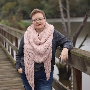 Op de afbeelding: Een vrouw draagt een roze gehaakte sjaal met een grijze trui en blauwe jeans. Ze staat op een houten brug.
