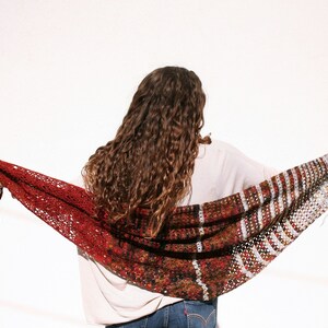 May include: A woman wearing a white shirt and jeans is holding a red and brown crocheted shawl. The shawl has a lacy pattern and is draped over her shoulders.