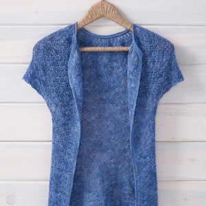 May include: A light blue crocheted cardigan with a delicate openwork pattern hangs on a wooden hanger against a white wood background.