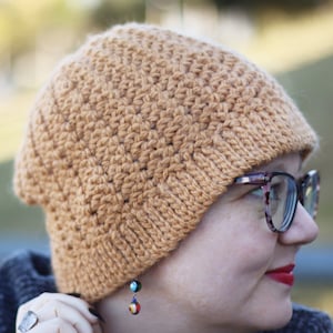 Op de afbeelding: Een close-up van een persoon die een bruine gebreide beanie draagt met een textuurpatroon. De beanie is naar beneden getrokken over hun voorhoofd en bedekt hun oren. De persoon draagt ook een bril en heeft een kleine oorbel in hun linker oor.