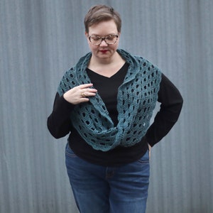 May include: A teal crocheted infinity scarf with a lacy pattern. The scarf is worn by a person with short hair and glasses, and they are wearing a black shirt and blue jeans.