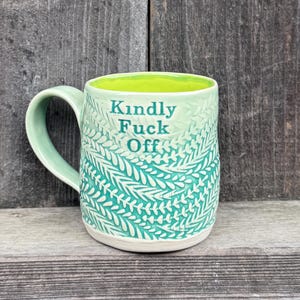 Puede incluir: Una taza de cerámica con exterior verde claro que presenta un patrón de hojas blancas texturizadas y el texto "Kindly FUCK Off" en verde oscuro. El interior de la taza es de color verde lima brillante e incluye un asa curva lateral.