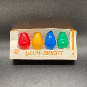 Puede incluir: Una caja con cuatro bombillas vintage GE Glow Bright en rojo, amarillo, azul y verde. Las bombillas tienen forma de lágrima y están en una caja de cartón con la inscripción "Glow Bright".