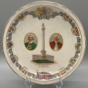 May include: A white ceramic plate with a gold rim and a floral design. The center of the plate features a monument with two portraits of men in military uniforms. The text "HERE DID WOLFE VICTORIOUS" is printed on the plate, along with "150th CENTENARY 1759 QUEBEC 1909".