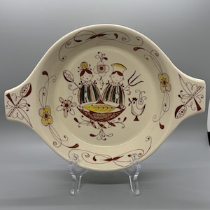 以下が含まれることがあります： クリーム色のヴィンテージサービング皿、持ち手付き。茶色、黄色、緑色の2つの人物、鳥、花のアクセントの気まぐれなイラストが特徴です。透明なスタンドに飾られています。