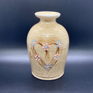 May include: A tan ceramic vase with a heart design painted in brown, pink, and blue. The heart is outlined in brown and filled with pink and blue dots.