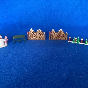 May include: A miniature Christmas village scene with a snowman and a child, a green bench, two stacks of firewood, and a row of small Christmas trees with red and white lights.