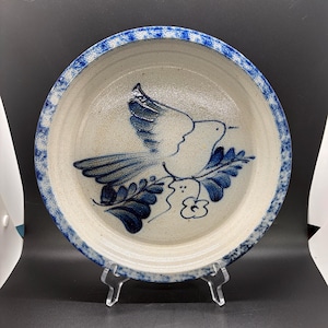 May include: A round ceramic pie dish with a speckled beige base and a blue sponge-painted rim. The centre features a hand-painted blue design of a bird in flight perched above leafy branches and a small flower, all rendered in a deep cobalt blue.