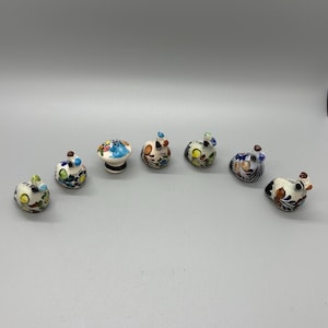 May include: A selection of seven small, hand-painted ceramic ornaments. The ornaments are mainly white with vibrant floral and abstract patterns in blue, yellow, orange, and brown. One ornament is mushroom-shaped, the others are animal-like.