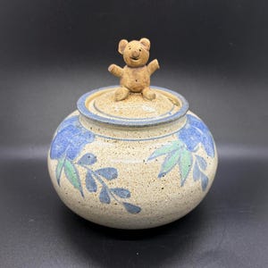 May include: A round ceramic jar with a speckled beige finish, featuring hand-painted blue flowers and green leaves. The lid is topped with a small, sculpted brown bear figure with outstretched arms, adding a tactile element to the piece.