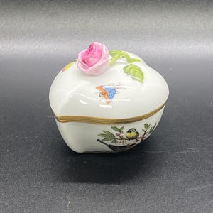 Könnte beinhalten: Weiße Porzellan-Schmuckschatulle mit einer rosa Rose oben. Die Schatulle hat einen goldenen Rand und ein gemaltes Design eines Vogels auf einem Ast. Auf der Seite ist auch ein Schmetterling gemalt. Dekorativer Artikel.