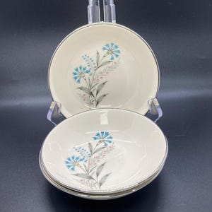May include: Set of four off-white ceramic bowls with a floral design. Each bowl features a silver rim and a delicate pattern of blue and pink flowers with gray stems. The bowls are stacked and displayed on a clear stand against a black background.