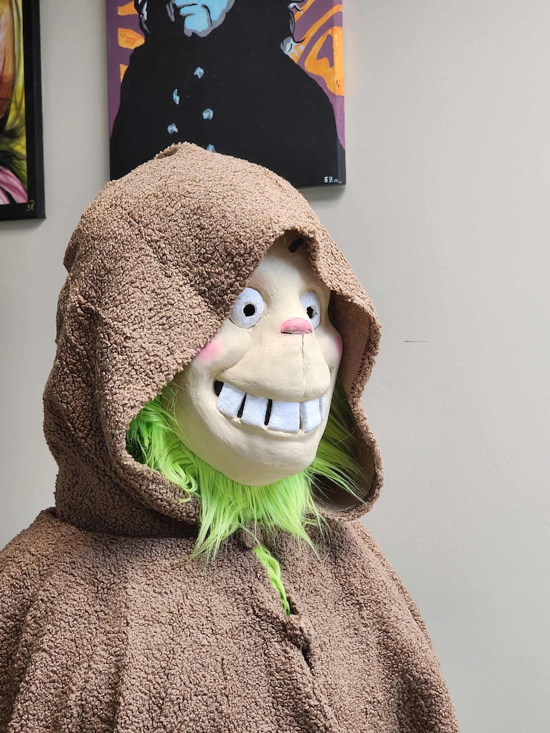 Op de afbeelding: Een bruin kostuum met capuchon en een lachend masker. Het masker heeft grote witte tanden, roze wangen en een groene baard. Het kostuum is gemaakt van een getextureerd, fuzzy materiaal.