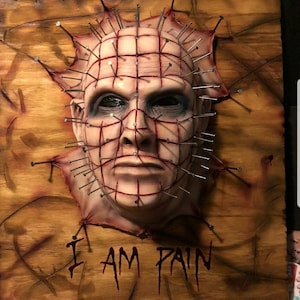 May include: A 3D horror mask of a face with a grid of nails and red lines. The mask is mounted on a wooden surface with the text "I AM PAIN" written below it.