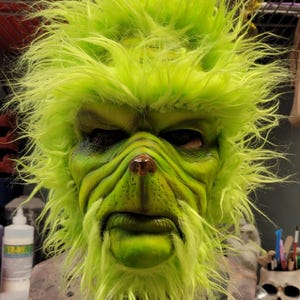 Puede incluir: Una máscara verde de Grinch con una cara peluda y pelo verde brillante. La máscara está hecha de un material realista y tiene una expresión detallada.
