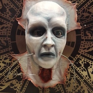 May include: A white face mask with a bloody, torn edge, attached to a black and brown patterned background. The mask has a distressed look and features a realistic depiction of a human face with piercing eyes and a slightly open mouth.