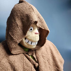Op de afbeelding: Een bruine cape met capuchon en bijpassend masker. Het masker heeft een cartoonachtig ontwerp met grote witte tanden, een roze neus en grote ogen. De cape is gemaakt van een getextureerd, fuzzy materiaal.