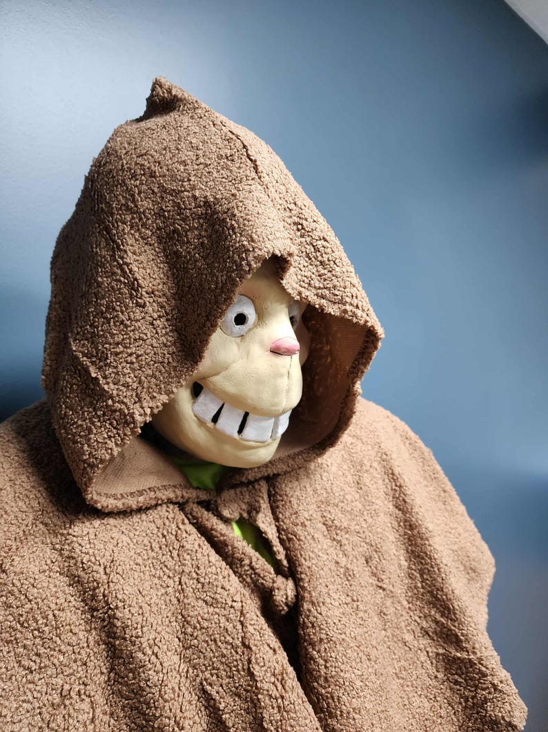 Op de afbeelding: Een bruine cape met capuchon en bijpassend masker. Het masker heeft een cartoonachtig ontwerp met grote witte tanden, een roze neus en grote ogen. De cape is gemaakt van een getextureerd, fuzzy materiaal.