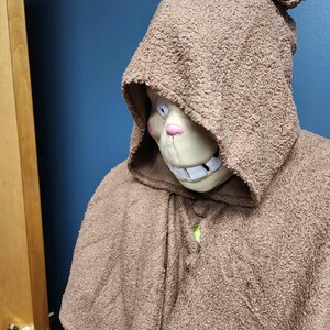 Op de afbeelding: Een bruine cape met capuchon met een getextureerde, fuzzy uitstraling. De capuchon omlijst een masker met een glimlachende uitdrukking, met witte tanden, een roze neus en een lichtbeige teint. De cape is bij de hals geknoopt.
