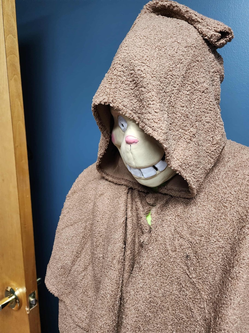 Op de afbeelding: Een bruine cape met capuchon met een getextureerde, fuzzy uitstraling. De capuchon omlijst een masker met een glimlachende uitdrukking, met witte tanden, een roze neus en een lichtbeige teint. De cape is bij de hals geknoopt.