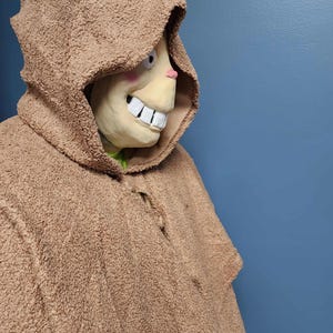 Op de afbeelding: Een bruin kostuum met capuchon en een masker met een grote, grijnzende grijns, roze wangen en een kleine roze neus. Het kostuum heeft een getextureerd, fuzzy uiterlijk. De achtergrond is effen blauw.