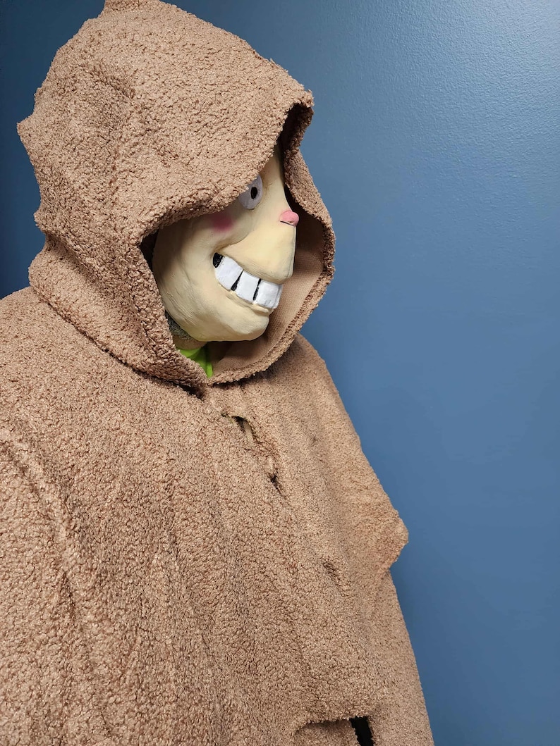 Op de afbeelding: Een bruin kostuum met capuchon en een masker met een grote, grijnzende grijns, roze wangen en een kleine roze neus. Het kostuum heeft een getextureerd, fuzzy uiterlijk. De achtergrond is effen blauw.