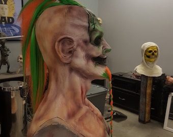 May include: A detailed, realistic, and grotesque-looking, hand-painted, latex mask of a zombie with a green and orange mohawk and a braided beard. The mask is mounted on a black tripod stand.