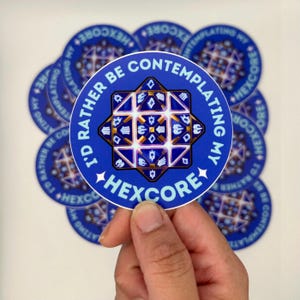 May include: A collection of blue round stickers with the text "I'd rather be contemplating my HEXCORE". Each sticker features a geometric design in white, gold, and blue. The stickers are held by a hand.