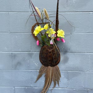 May include: A decorative wall hanging featuring a turtle shell-shaped container filled with yellow and pink flowers, accented with brown feathers and tall grasses. The arrangement is mounted on a gray brick wall, creating a rustic, natural aesthetic.