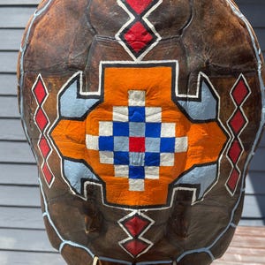 May include: A painted turtle shell with a geometric design in orange, blue, red, and white. The shell is brown and has a beaded fringe hanging from the bottom.