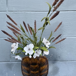 May include: A decorative wall-mounted vase made from a turtle shell, filled with white flowers, green leaves, and brown wheat stalks. The vase is a dark brown color with a textured surface, and the flowers are a bright white.