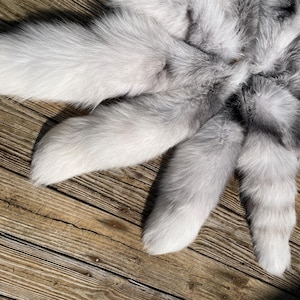 May include: A circular arrangement of fluffy, white and gray fur tails. The tails radiate outwards from a central point, with a mix of white and gray fur. The tails are arranged on a wooden surface.