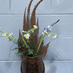 May include: A decorative wall hanging featuring a turtle shell planter filled with flowers and pheasant feathers. The shell is a dark brown color, and the feathers are brown with black markings. The flowers are white, yellow, and purple.