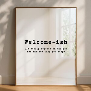 Puede incluir: Una lámina decorativa rectangular en un marco metálico fino con el texto negro estilo máquina de escribir Welcome-ish seguido de la frase entre paréntesis It really depends on who you are and how long you stay sobre fondo blanco.