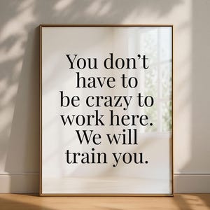 May include: A rectangular wall art print in a thin metallic frame featuring black serif typography on a white background. The text reads: You don't have to be crazy to work here. We will train you. The design is centered within the frame.