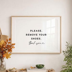 May include: A horizontal wall art print in a light wood frame featuring the black text: PLEASE. REMOVE YOUR SHOES. Thank you. The print is displayed above a wooden surface with a glass vase of autumn leaves, a small bowl, and books.