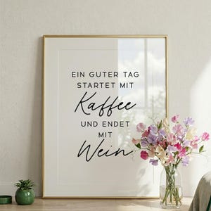 May include: A framed print with the text "EIN GUTER TAG STARTET MIT Kaffee UND ENDET MIT Wein" in black script. The frame is gold. A vase of colorful flowers sits to the right of the frame, and a small potted plant is on the left.