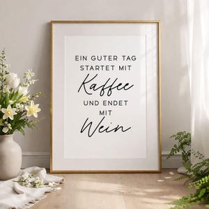 May include: A framed print with a gold border and white mat. The print features black script that reads, "EIN GUTER TAG STARTET MIT Kaffee UND ENDET MIT Wein." A vase of white flowers sits to the left.
