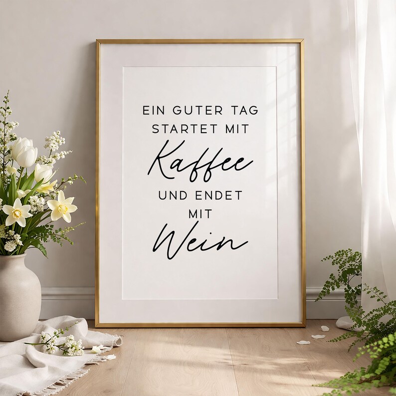 May include: A framed print with a gold border and white mat. The print features black script that reads, "EIN GUTER TAG STARTET MIT Kaffee UND ENDET MIT Wein." A vase of white flowers sits to the left.