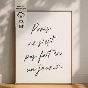 Könnte beinhalten: Gerahmter Kunstdruck mit dem Satz "Paris ne s'est pas fait en un jour" in eleganter schwarzer Schrift. Der Druck hat einen cremefarbenen Hintergrund und einen goldenen Rahmen. In der oberen linken Ecke befindet sich ein "DIGITAL PRODUCT"-Label mit Cloud- und Drucker-Symbolen.