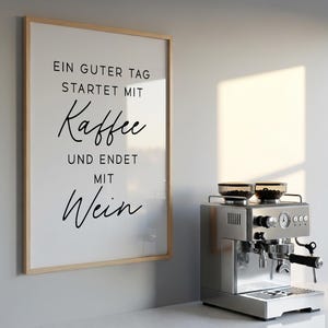 May include: A framed print with the text "EIN GUTER TAG STARTET MIT Kaffee UND ENDET MIT Wein" hangs next to a stainless steel espresso machine. The machine has a portafilter and two glass containers on top.