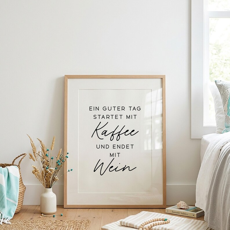 May include: A framed print with the text "EIN GUTER TAG STARTET MIT Kaffee UND ENDET MIT Wein" in black script. The print is in a light wood frame and is leaning against a white wall. A vase of dried flowers and a woven basket are in the foreground.