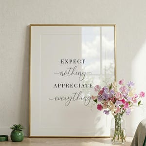 Può includere: Una stampa incorniciata con le parole "EXPECT nothing APPRECIATE everything" in elegante scrittura. La cornice dorata circonda uno sfondo bianco. Un vaso di fiori colorati è posto accanto all'opera d'arte incorniciata, con una piccola pianta in vaso in primo piano.