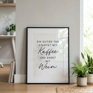 May include: Framed print with the text "EIN GUTER TAG STARTET MIT Kaffee UND ENDET MIT Wein" in black script on a white background. The frame is black. The print is displayed on a wooden floor.
